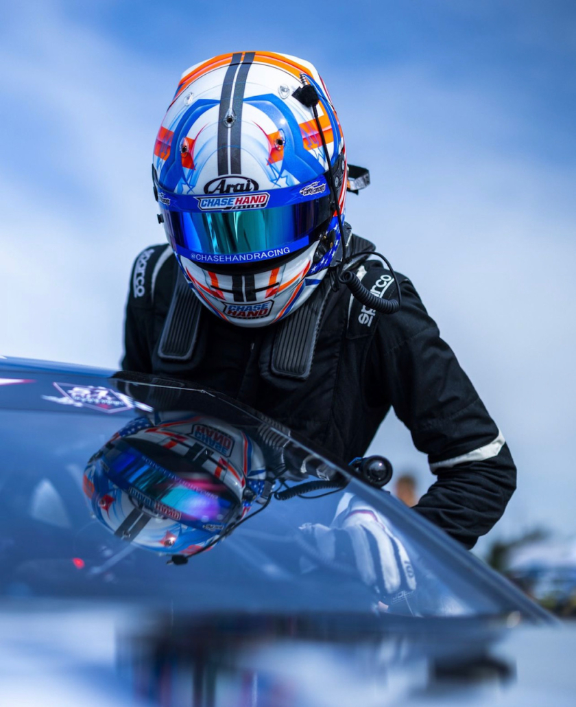 In a reflective helmet and racing gear, Chase leans over the front windshield of a black race car.