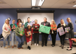 A group of Visions staff in festive holiday clothes stands in the front of Visions' board room holding wrapped gifts.