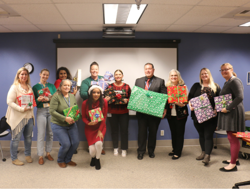A group of Visions staff in festive holiday clothes stands in the front of Visions' board room holding wrapped gifts.