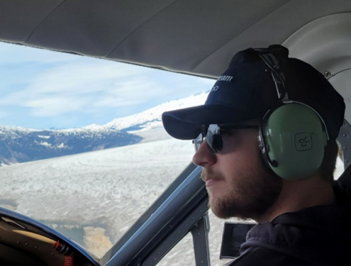 In a navy cap, black sunglasses and green over-the-ear headset, Hunter sits in the cockpit of a plane flying over clouds and looking out over the horizon.