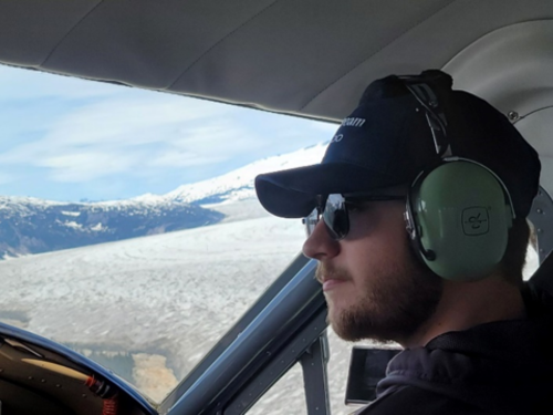 In a navy cap, black sunglasses and green over-the-ear headset, Hunter sits in the cockpit of a plane flying over clouds and looking out over the horizon.