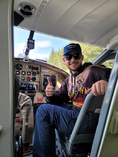 In a navy cap, black sunglasses and multi-colored sweatshirt, Hunter sits in the cockpit of a plane and gives a "thumbs up" to the camera.