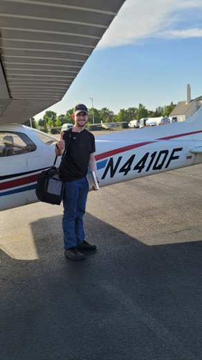 In a cap, glasses, a black shirt and jeans, Hunter stands next to a plane on the runway with his gear and a book in his hand, giving a thumbs up to the camera.