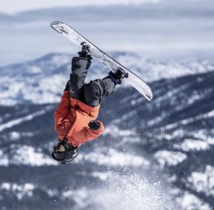 In black snow pants, a red jacket and black helmet and goggles, Jax does a flip on his snowboard with the view of mountains in the background.