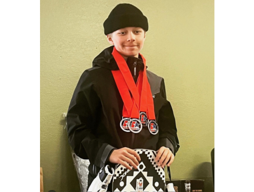 Jax Allen poses with his snowboard wearing a black beanie, black jacket, and multiple red medals around his neck.