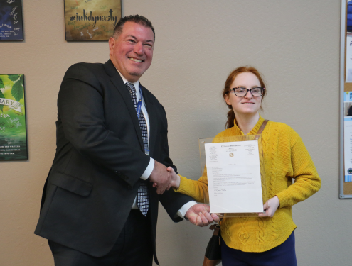 Superintendent Olmos stands next to Tori Berg in the Learning Center, holding the framed letter from Senator Niello in one hand and shaking Tori's hand with the other.