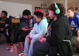 A group of students all sit around a projector screen and watch as one student plays the video game Super Smash Bros.