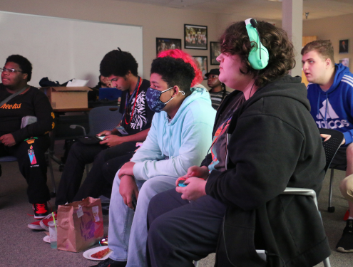 A group of students all sit around a projector screen and watch as one student plays the video game Super Smash Bros.