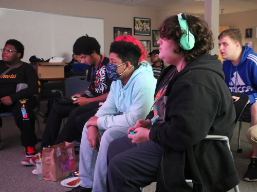 A group of students all sit around a projector screen and watch as one student plays the video game Super Smash Bros.