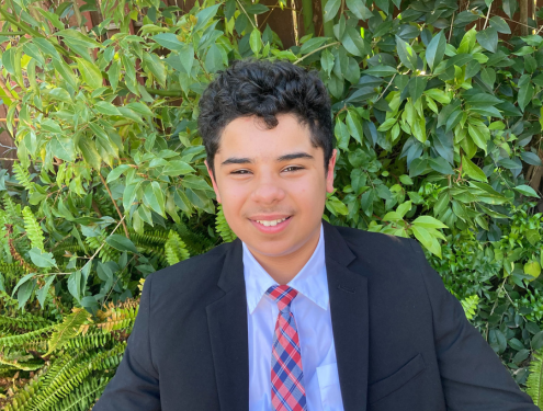 In a black suit, light purple shirt and red and blue tie, Bassam poses for a photo in front of a bright green bush.