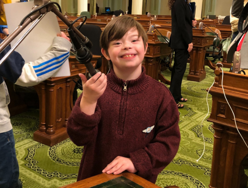 In a maroon-colored sweater, Dominic smiles for the camera as he holds a podium microphone at the California State Capitol.