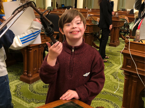 In a maroon-colored sweater, Dominic smiles for the camera as he holds a podium microphone at the California State Capitol.