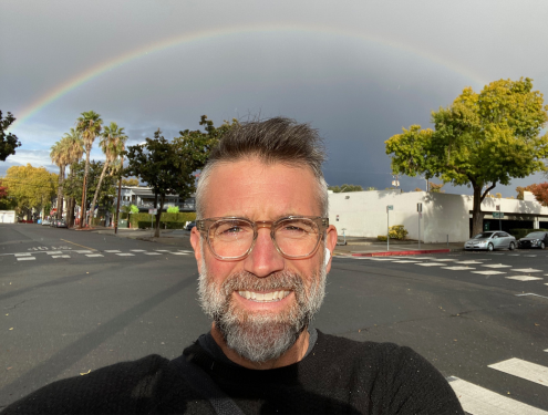 Mitch takes a selfie wearing a black t-shirt and Apple Airpods standing underneath a rainbow.