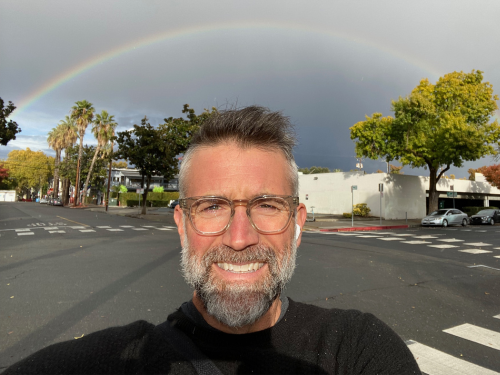Mitch takes a selfie wearing a black t-shirt and Apple Airpods standing underneath a rainbow.