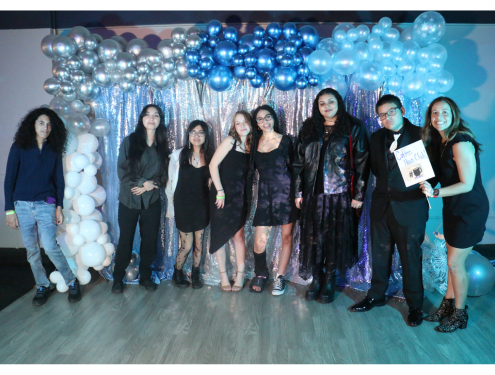 Visions' Coffee House Hangout club gathers together in front of a gray, white, and blue backdrop of streamers and balloons and takes a group photo with their club advisor holding a sign that says "Coffee House Club".