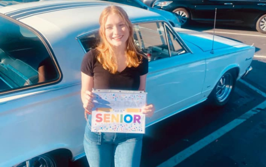 Visions student standing in front of a blue car holding a small sign that says "Senior"