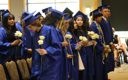 Visions graduates in caps and gowns at graduation standing up holding white roses
