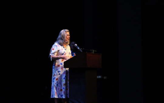 Alumni Hall of Fame Inductee Jessica Chavez's teacher speaking on her behalf on stage at the August staff back to school event.