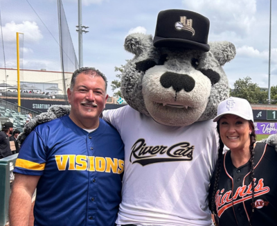 Visions Superintendent Dr. Steve Olmos wears a custom blue and yellow Visions baseball jersey and stands next to the Sacramento River Cats mascot Dinger and Visions staff Lisa Adams.
