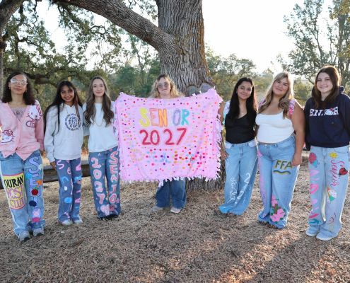A group of students in custom decorated jeans and a blanket for senior year gather underneath a tree for a group photo.