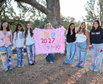 A group of students in custom decorated jeans and a blanket for senior year gather underneath a tree for a group photo.
