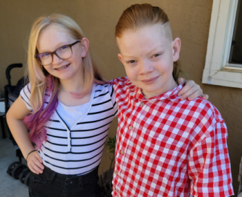 Two young siblings, a girl with glasses and a black and white striped shirt and a boy in a red and white checkered shirt, stand next to one another and smile for a first day of school photo.
