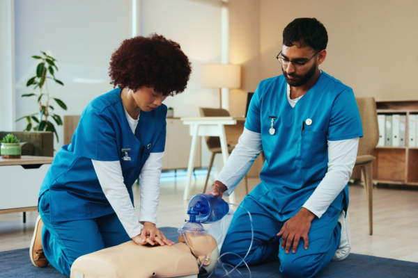 Male and female students in blue scrubs kneeling over a medical mannequin on the ground practicing CPR