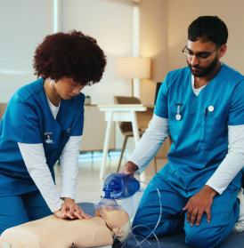 Male and female students in blue scrubs kneeling over a medical mannequin on the ground practicing CPR