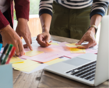 Close up photo of two people working together with sticky notes, pens, and a laptop.