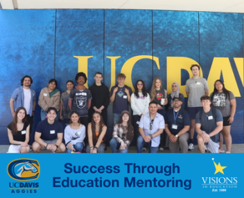 A group of Visions high school students gather for a group photo in front of a blue and yellow wall that says "UC Davis."