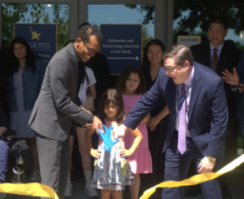 Two Board members cut gold ribbon with large blue scissors at ceremony.